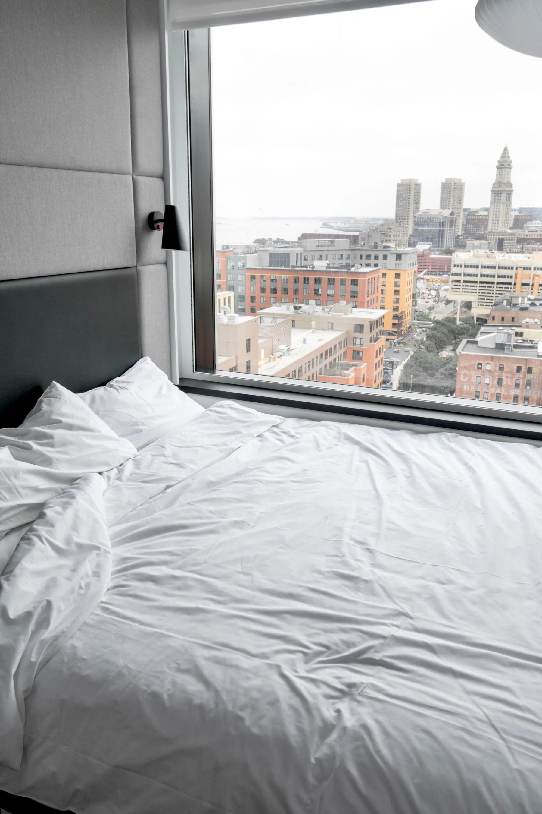 Cama feita com roupa branca lavada, num quarto com vista para a cidade
